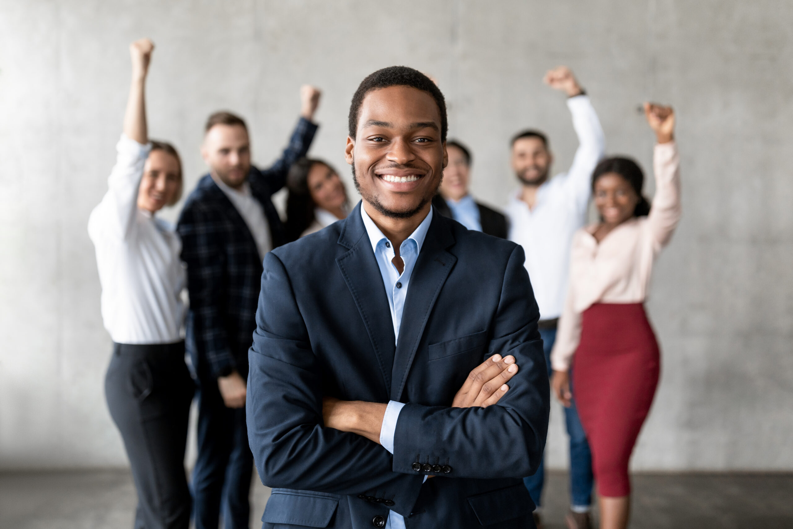 A confident professional standing with arms crossed while a motivated team celebrates behind him, representing strong leadership and management.